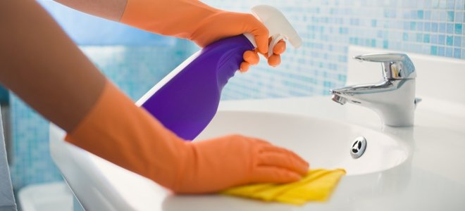 Manos enguantadas limpiando un fregadero con una botella de spray morada y un paño amarillo