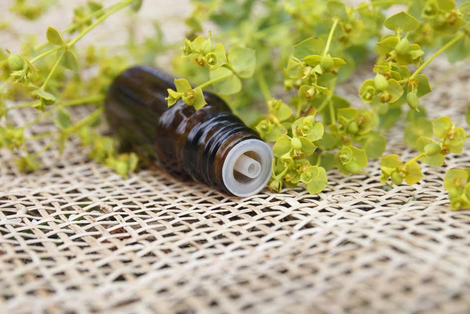 Una botella de aceite esencial marrón sobre arpillera con hierbas al lado. 