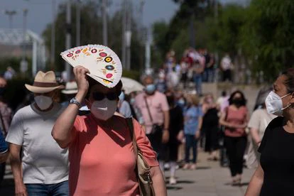 Una mujer se protege del sol y el calor con un abanico en la cola que da acceso al centro de vacunación instalado en el Estadio Olímpico de Sevilla.