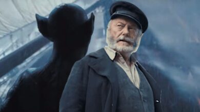 Liam Cunningham in The Last Voyage of the Demeter in a captain's uniform and thick beard, backdropped by a shadowy image of a monstrous Dracula with rat-like ears gazing down upon a ship at sea, waves crashing onto the deck