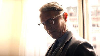 Mads Mikkelsen as Jurgen Voller in Indiana Jones 5 wearing glasses and a suit, looking menacing against a curtained window