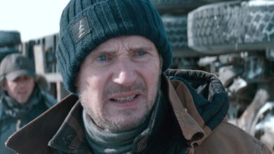 Liam Neeson as Mike McCann in The Ice Road grimacing intensely in the cold with a crashed truck behind him