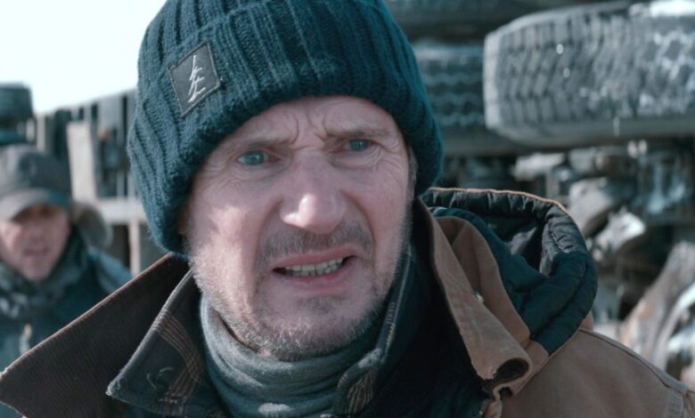 Liam Neeson as Mike McCann in The Ice Road grimacing intensely in the cold with a crashed truck behind him