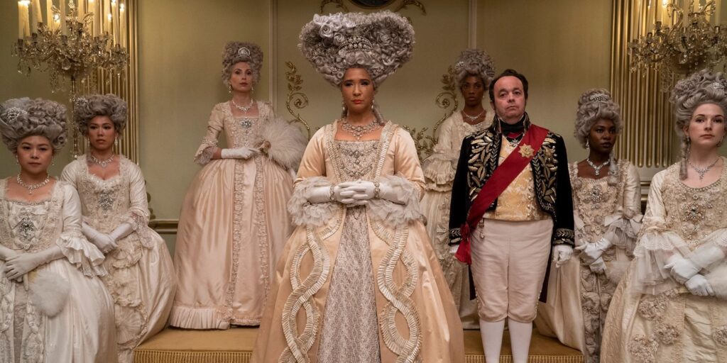 Queen Charlotte in Queen Charlotte: A Bridgerton Story at court surrounded by women in white dresses with a male court servant next to her.