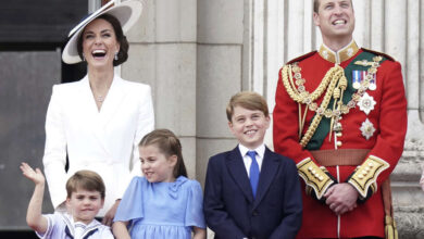 Catherine, la princesa de Gales, sonríe ampliamente con un traje blanco y un sombrero blanco de gran tamaño junto a su esposo, el príncipe William, vestido de militar. Los tres hijos de la pareja, Louis, Charlotte y George, están de pie frente a ellos, también sonrientes y vestidos de gala. Louis parece estar en la mitad de la ola.