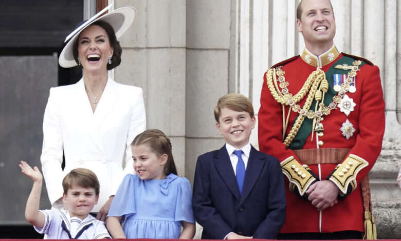 Catherine, la princesa de Gales, sonríe ampliamente con un traje blanco y un sombrero blanco de gran tamaño junto a su esposo, el príncipe William, vestido de militar. Los tres hijos de la pareja, Louis, Charlotte y George, están de pie frente a ellos, también sonrientes y vestidos de gala. Louis parece estar en la mitad de la ola.