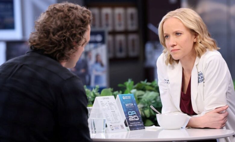Hannah Asher and Sean Archer sit at a table in the hospital in Chicago Med season 8, episode 20. Asher wears a white doctor's coat over maroon scrubs. Sean wears a dark blue plaid shirt.