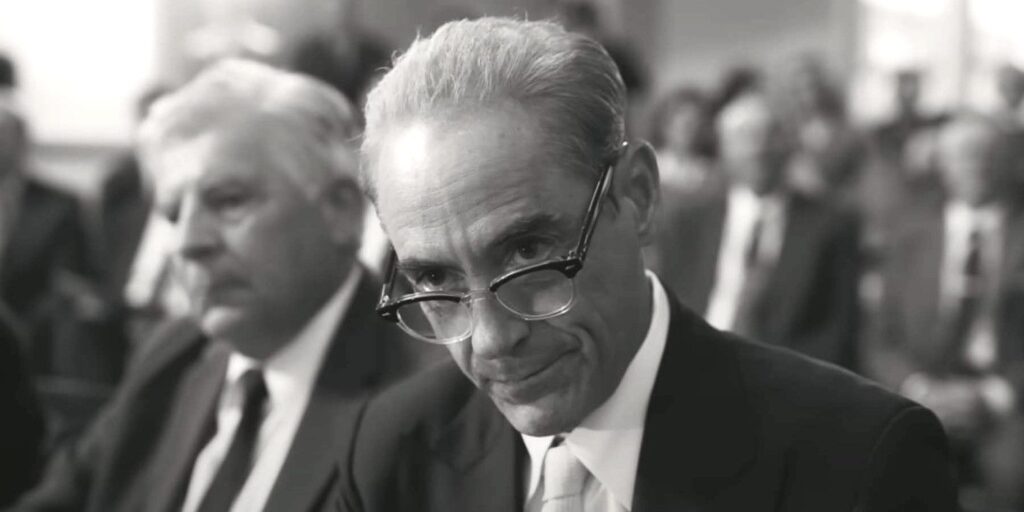 Robert Downey Jr in Oppenheimer wearing '40s period attire in a large meeting room, leaning forward to peer over his horn-rimmed glasses