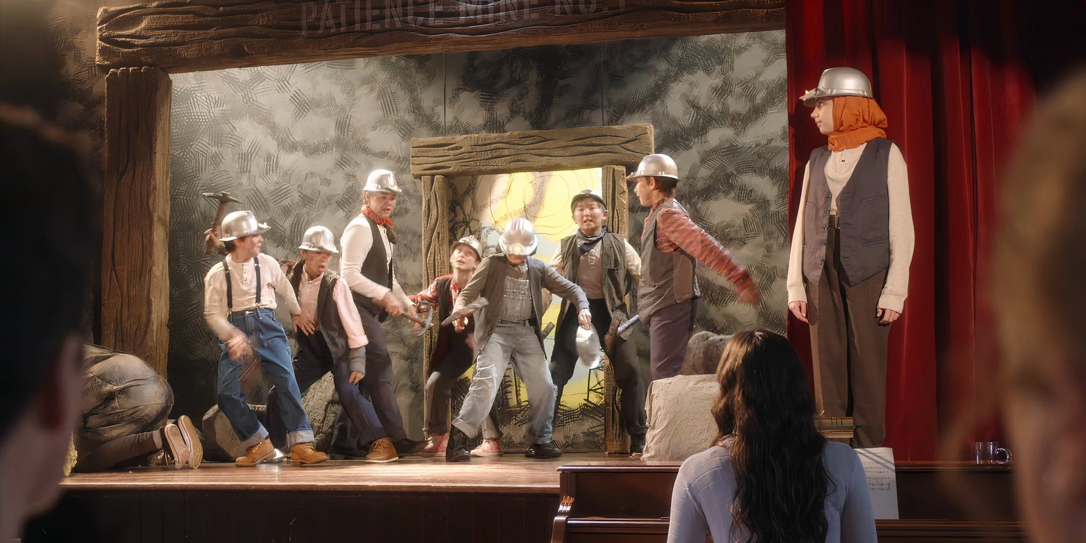 Niños con trajes mineros que actúan en un escenario con Sahar parado a un lado frente a una cortina roja