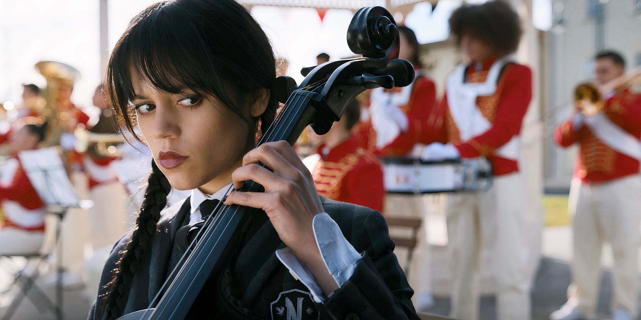 Jenna Ortega como el miércoles tocando el violonchelo frente a una banda de música, con los ojos mirando hacia abajo y hacia la derecha