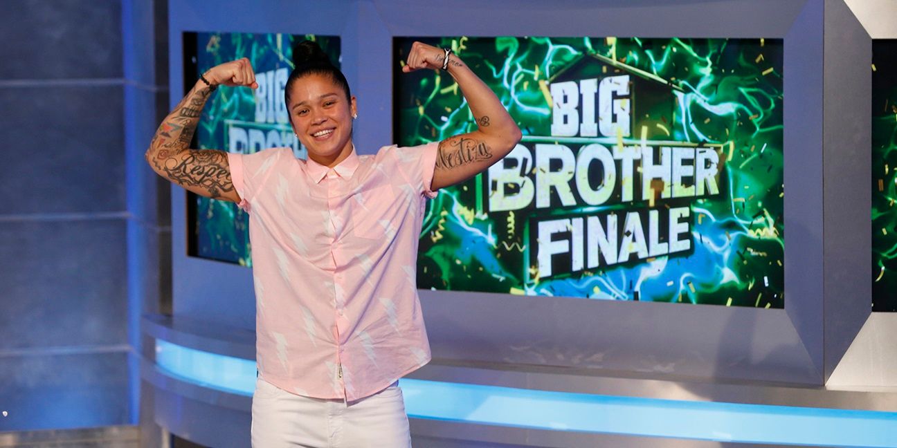Kaycee flexionando sus músculos en el Finale Big Brother con una pantalla detrás de ella que muestra "Big Brother Finale" y el logotipo de TheW. Ella lleva pantalones blancos y una camisa de manga corta con botones rosa.