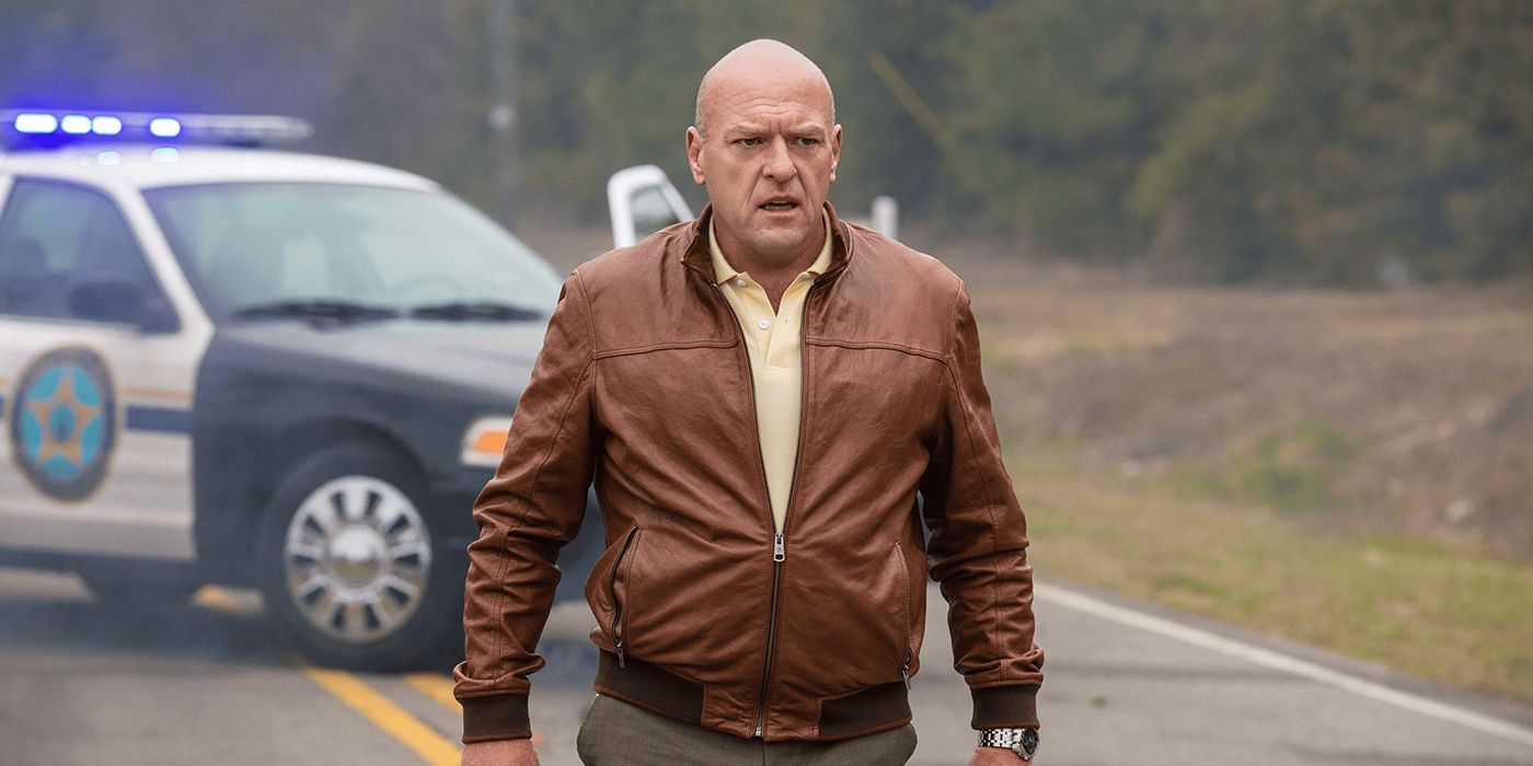 Dean Norris como Big Jim parado en la calle, mirando preocupado por un auto de la policía detrás de él, en debajo del domo.