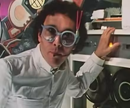 Una persona tiene una camisa blanca, gafas blancas y cabello de longitud media. Está de pie con una mano sobre una radio. Detrás de él hay una pared pintada de color púrpura con toques de color.