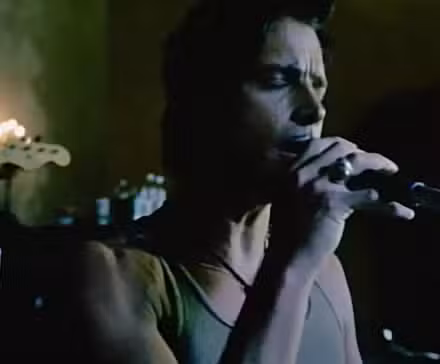 El cantante de Audioslave tiene cabello negro y bigote fino. Lleva una camiseta gris y se lleva un micrófono a la boca. La pared detrás de él es verde y se ven parte de un bajo y una batería.