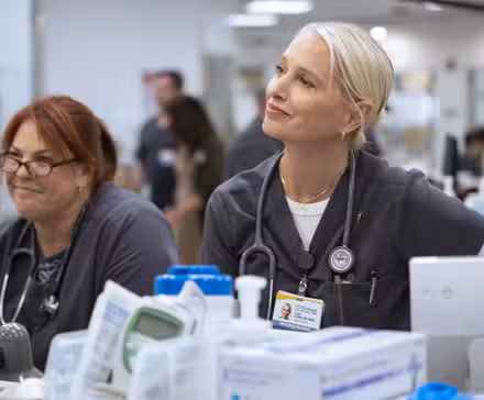 Lena (Lesley Boone) y Dana (Katherine LaNasa) sonriendo en el escritorio de la enfermera en la temporada 2 de The Pitt