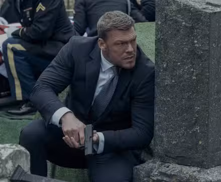 Reacher acurrucado detrás de una lápida con una pistola en la temporada 2 de Reacher