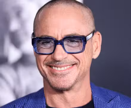 Robert Downey Jr., con la cabeza rapada, usa elegantes gafas azules y sonríe mientras asiste a un evento