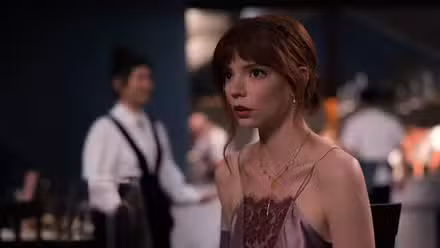 Anya Taylor Joy como Margot en The Menu con un vestido, luciendo aterrorizada, con los camareros del restaurante al fondo.