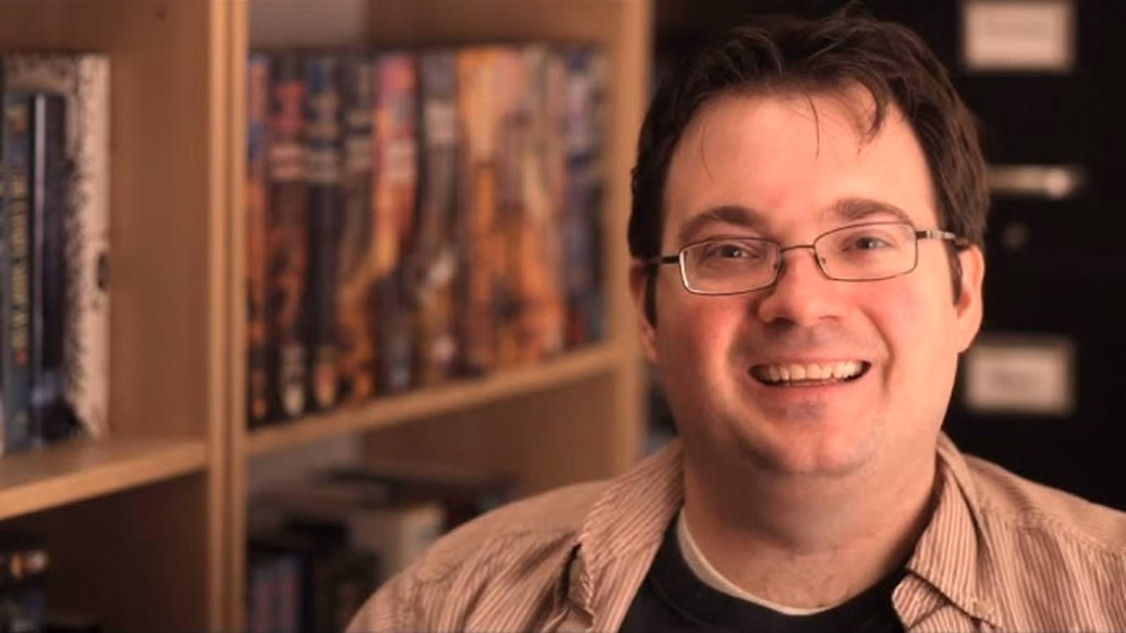 Brandon Sanderson (primer plano) con sus libros en un estante (al fondo)